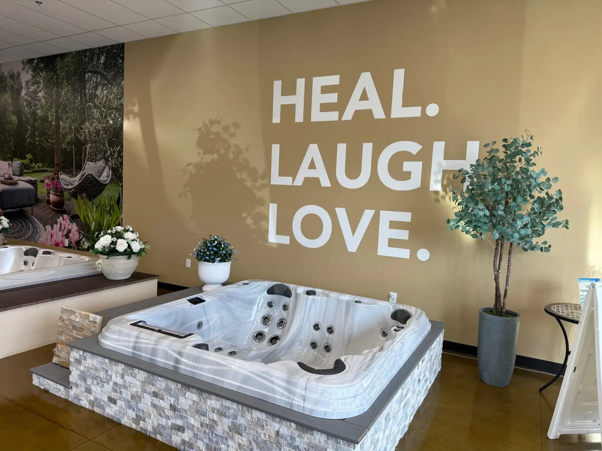 Hot tub display in Creative Hot Tub Designs showroom in Reno, NV, featuring "HEAL. LAUGH. LOVE." wall decor, surrounded by potted plants and floral arrangements.