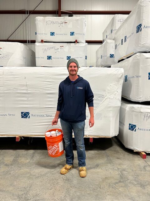 Matt, Water Care Technician at Creative Hot Tub Designs, holding a bucket of water care supplies in a warehouse filled with packaged Artesian Spas, Sparks NV.