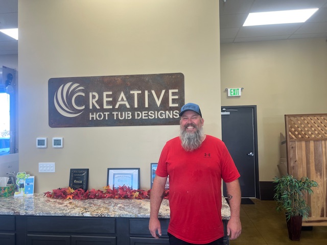 Smiling man in red shirt standing in front of Creative Hot Tub Designs sign, showcasing the welcoming environment of the Reno, NV location.