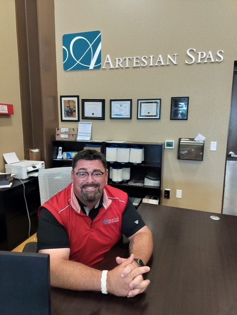 Tommy, a sales consultant at Creative Hot Tub Designs, seated at his desk in Sparks, NV, with an Artesian Spas sign in the background, showcasing a welcoming customer service environment.