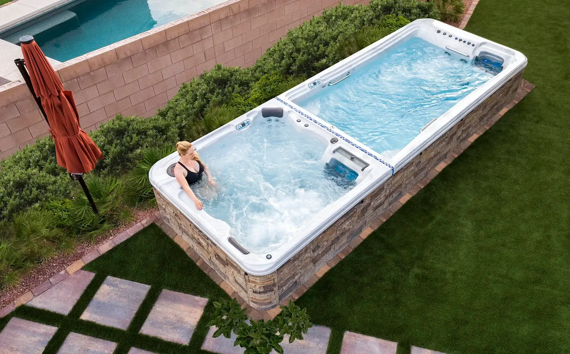 Swim spa with lounge seating and jets, featuring a woman enjoying the water, surrounded by greenery and patio stones, from Creative Hot Tub Designs in Reno, NV.