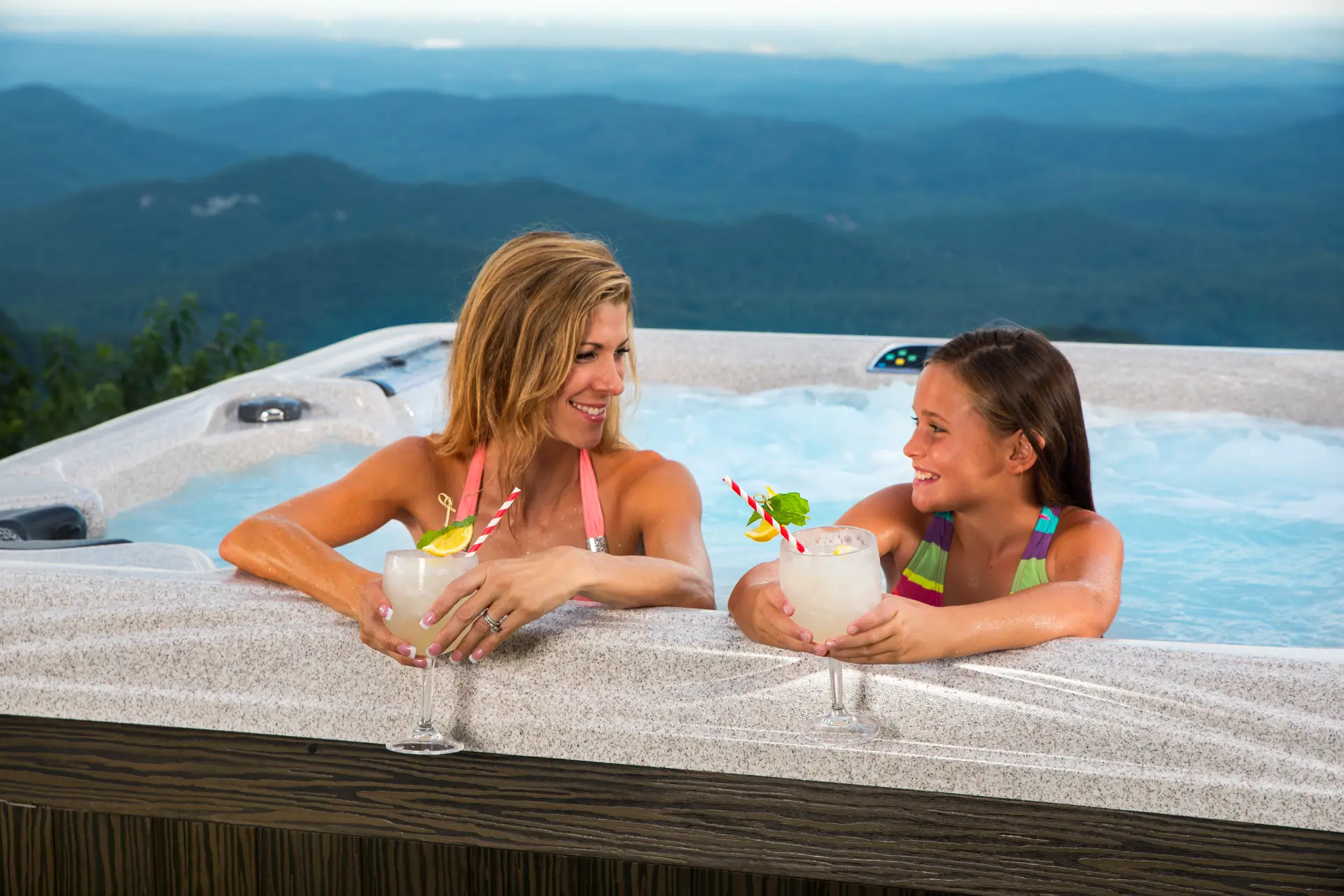 Woman and girl enjoying cocktails in hot tub with scenic mountain view, promoting family relaxation and outdoor living, relevant to Creative Hot Tub Designs in Reno NV and Sparks NV.